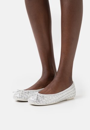 Jambes à peau foncée portant des ballerines argentées tissées avec de petits nœuds à l'avant, debout sur une surface blanche unie.