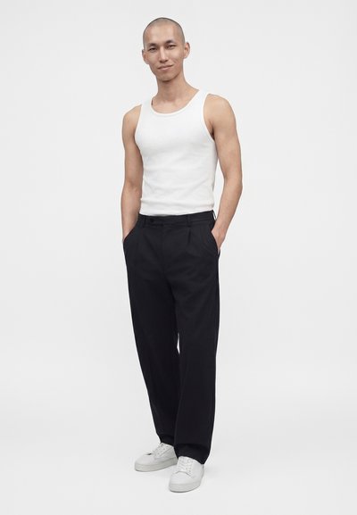 Homme portant un débardeur blanc, un pantalon noir à jambes larges et des baskets blanches, debout les mains dans les poches sur fond blanc.
