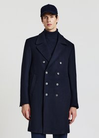 Cappotto doppiopetto in lana blu navy con revers appuntito, sei bottoni d'argento e tasche laterali, indossato sopra un dolcevita a maglia scura. Cappello abbinato.