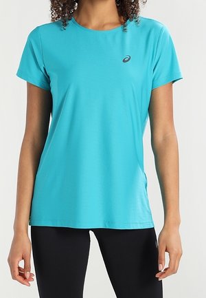 Vrouw draagt een turquoise korte mouw sportshirt en zwarte leggings, staand met ontspannen armen langs haar zij.