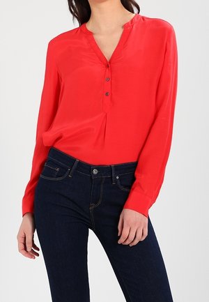 Kobieta ubrana w jasnoczerwoną bluzkę z długim rękawem, dekoltem w serek i guzikami, w połączeniu z dopasowanymi ciemnoniebieskimi dżinsami.