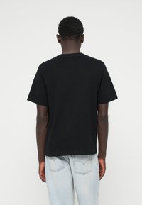 Must mustaga T-särk lühikeste varrukatega, ümar kaelus ja vabalt langev lõige. Seljas helesinised denim teksad, vaadatud selja tagant.