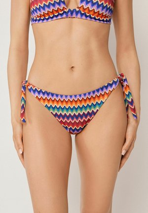 Femme portant un bas de bikini coloré à motif zigzag avec des liens sur les côtés et un haut de bikini assorti, visible du ventre aux cuisses.