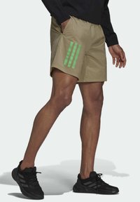 Beiga träningsshorts med tre gröna vertikala ränder på vänster sida, tillverkade av lätt material, med ett elastiskt midjeband och fickor.