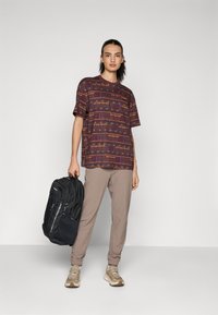Graphic patterned short-sleeve shirt in burgundy, paired with beige tapered pants and gray sneakers. Holds a black backpack.