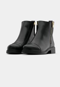 Paire de bottines en cuir noir avec talons bas et fermetures éclair dorées sur les côtés extérieurs, sur fond blanc uni.
