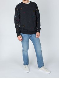 Uomo che indossa un felpa scura con motivi di uccelli multicolori, jeans azzurri chiari e sneakers bianche, in piedi contro uno sfondo neutro.