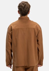 Braune, übergroße Jacke mit einem schlichten Design, Knopfdetail an den Ärmeln und einem glatten, strukturierten Stoff. Ansicht von hinten.