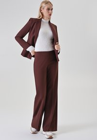 Bruine op maat gemaakte blazer met bijpassende broek met wijde pijpen, gelaagd over een gestructureerd wit trui. Kenmerkt zich door een gladde stof en een slim fit.