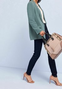 Veste blazer vert clair, haut crème, jean foncé, et sandales à talons roses. Porte un sac à main beige et effet peau de serpent avec des accents noirs.