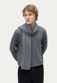 En grå ullblandad scarf hänger löst runt halsen, tillsammans med en matchande grå tröja. Mjuk textur och enkel, modern design.