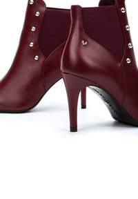 Botas de tobillo de charol burdeos con tacón alto, confeccionadas en cuero suave, que presentan paneles laterales elásticos y tachuelas decorativas en tono dorado.