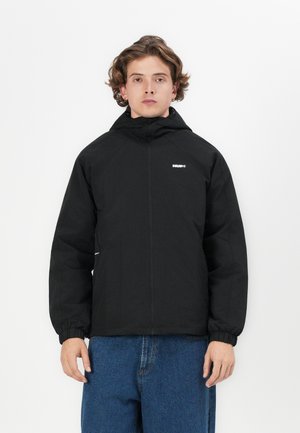 Veste à capuche noire avec des poignets élastiques et une fermeture éclair à l'avant. Présente un logo sur la poitrine et une coupe décontractée. Portée avec un pantalon en denim bleu.