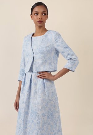 Frau trägt ein hellblaues, geblümtes Jacquardkleid mit einer passenden kurzen Jacke, posiert mit einer Hand in der Hüfte vor einem einfarbigen Hintergrund.