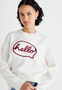 Sweat-shirt blanc avec une bulle de dialogue rouge brodée portant le mot "bonjour". Manchons et encolure côtelés, texture de tissu douce.