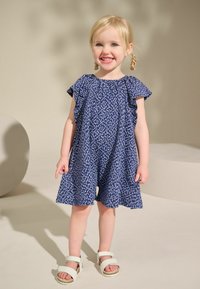 Robe bleue à motifs avec des manches volantes, un col rond et une coupe décontractée. Associée à des sandales blanches à lanières. Fond neutre.