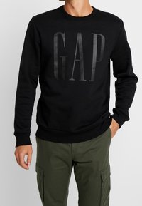 Sweat-shirt noir avec logo "GAP" en gris foncé, col rond, manches longues et poignets côtelés. Porté avec un pantalon cargo vert olive.