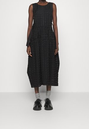 Femme portant une robe midi noire texturée sans manches avec des liens latéraux, des chaussettes grises et des chaussures noires à lacets, debout devant un fond uni.