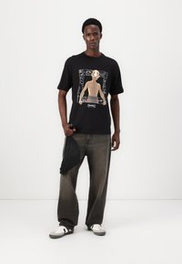 Sort bomulds T-shirt med grafisk design af en karakter i en rektangulær ramme. Sammen med løse grå jeans og grå sneakers.