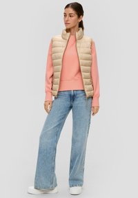Beige gewatteerde vest over een roze trui, gecombineerd met wijde lichtblauwe jeans en witte chunky sneakers. Zachte texturen, casual pasvorm.