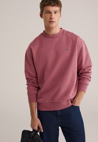 Rdečkasto roza majica z okroglim izrezom, rebrastimi manšetami in majhnim vezenim logotipom na prsih, kombinirana s temnimi denim kavbojkami.