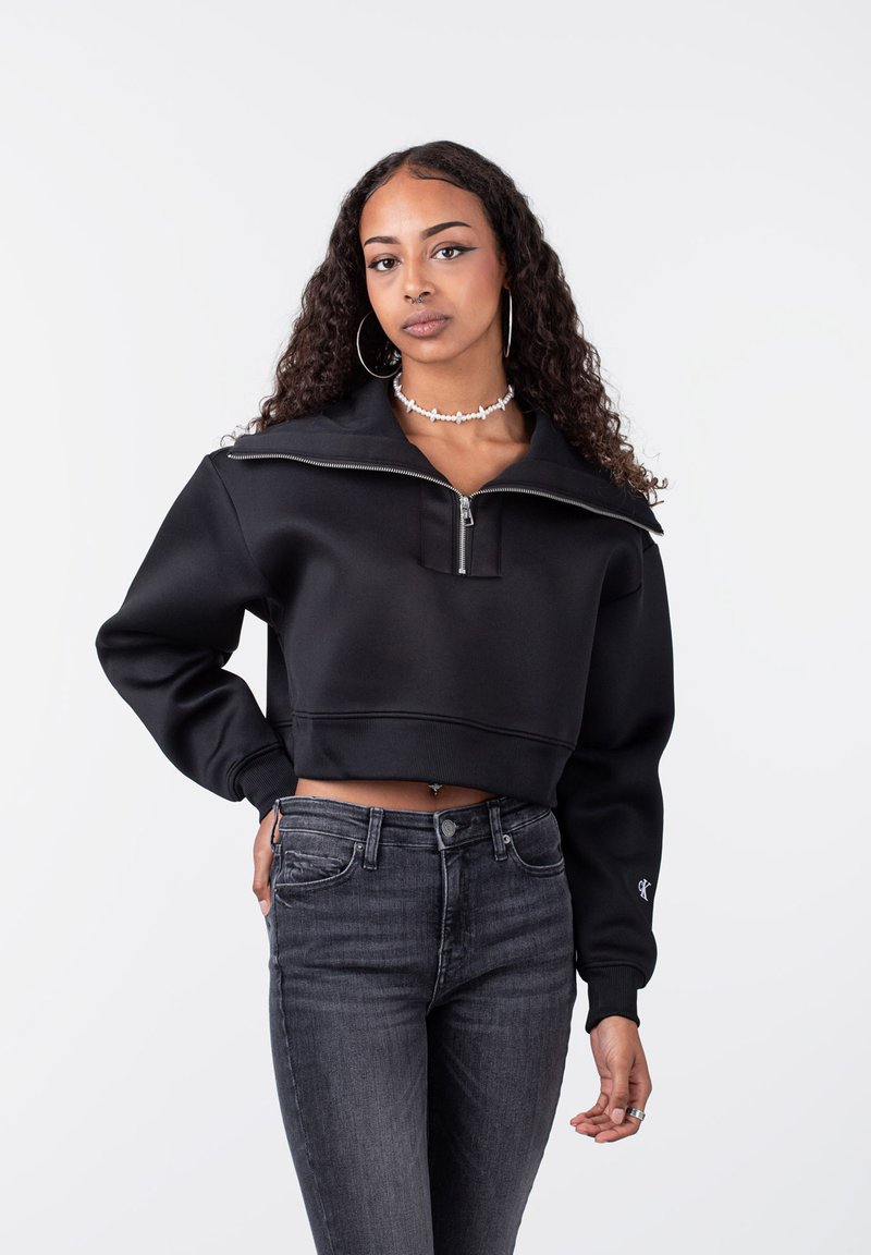 Sudadera negra corta con un cuello alto y cierre de media cremallera. Cuenta con mangas largas y un dobladillo acanalado, combinada con unos jeans oscuros ajustados.