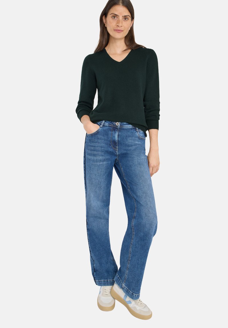 Dunkelgrüner V-Ausschnitt-Pullover, hellblaue gerade Jeans und weiße Sneakers mit blauen Akzenten. Baumwollmischung, lockere Passform und klassisches Design.