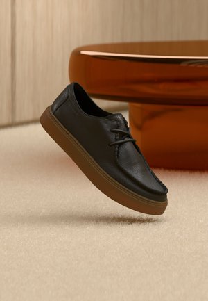 Zapato casual de cuero negro con suela marrón flotando sobre una alfombra beige, con una mesa redonda marrón brillante al fondo.