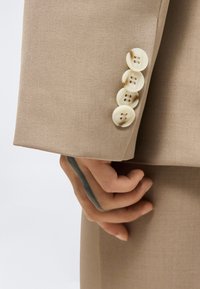 Μπεζ μανίκι σακακιού με τέσσερα ανοιχτόχρωμα κουμπιά, μερικώς ορατό χέρι από κάτω με σκούρο γραμμικό τατουάζ στον καρπό.