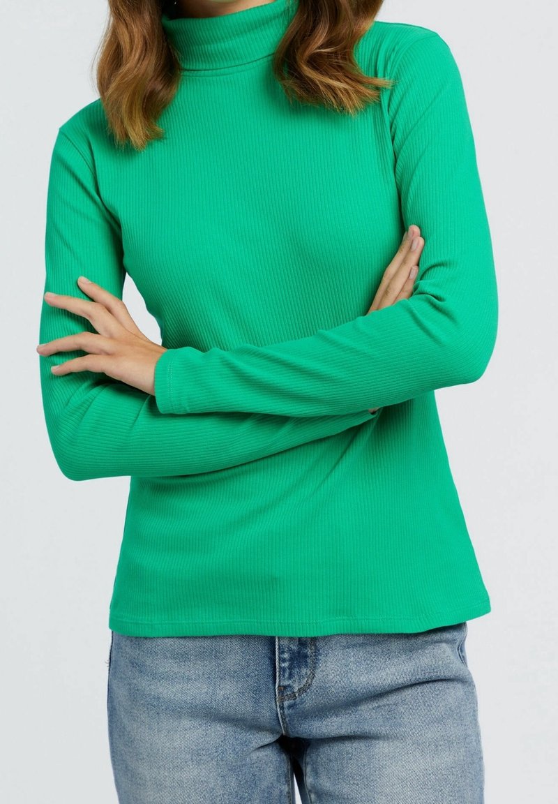 Femme en col roulé côtelé vert, les bras croisés, portant un jean bleu clair, vue du cou aux hanches sur fond uni.