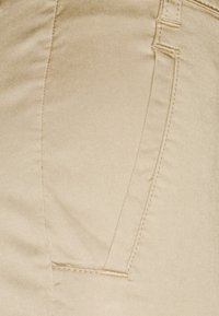 Tissu beige avec une texture lisse et un détail de poche cousue, présentant des lignes épurées et une coupe ajustée.