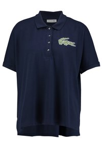 Námořnická modrá polo tričko z bavlny, má krátké rukávy, zapínání na tři knoflíky a zelené logo krokodýla Lacoste na levé straně hrudi.