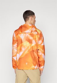 Lysorange jakke med et geometrisk mønster, hætte, elastiske manchetter og et logo på ryggen. Letvægts, vandafvisende materiale.