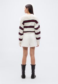 Λευκό κοντό πουλόβερ με κόκκινες και ναυτικές οριζόντιες ρίγες, πλεκτή υφή με χονδρή πλέξη· συνοδεύεται από λευκό σορτς και μαύρες μπότες μέχρι το γόνατο.