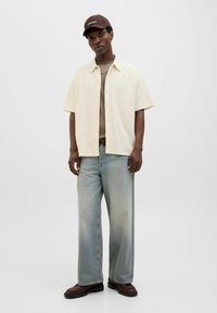 Camisa de botones de manga corta crema sobre una camiseta beige, combinada con vaqueros de pierna ancha azul claro y calzado oscuro, con una gorra marrón.