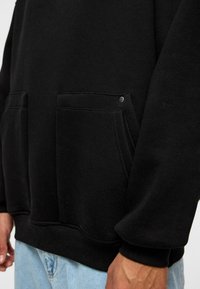 Zwarte sweatshirt met een zachte textuur, voorzien van twee voorzakken, zichtbare stiksels en een ronde zoom. Subtiele metalen accenten bij de zakken.