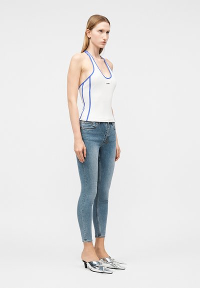 Γυναίκα με καστανόξανθα ίσια μαλλιά, που φοράει λευκό αμάνικο τοπ με μπλε περιθώριο, ανοιχτό μπλε στενά τζιν και ασημί μυτερά mule με τακούνι.