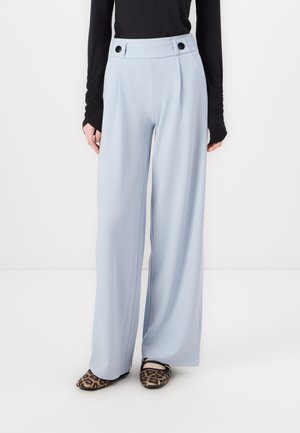 Pantalons larges bleu clair avec des plis, présentant une taille haute et deux boutons noirs. Tissu lisse, associé à des chaussures plates imprimées.