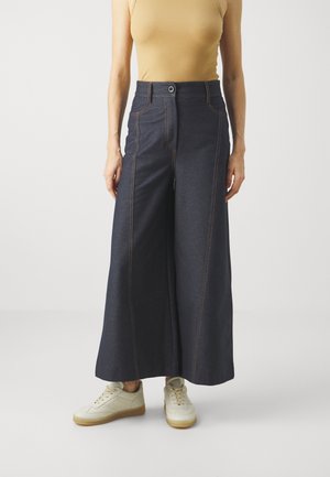 Kvinde iført mørke, vidde vide, højtaljede denimbukser med knapper foran og beige sneakers, stående mod en ensfarvet lys baggrund.