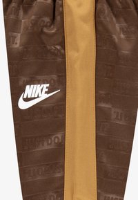 Rjavi športni hlače s svetlo rjavim pasom, ki imajo teksturiran vzorec in bel logotip Nike na spodnjem delu noge.