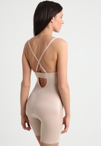 Beige corrigerend bodysuit met verstelbare kruisbanden en een uitgesneden rug. Gladde textuur, knielang ontwerp met een onderbroekstijl onderkant.