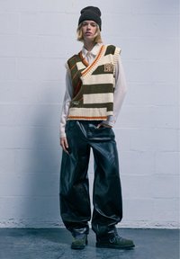 Strickweste mit wechselnden Streifen in Creme, Olivgrün und Rostorange; lockere schwarze Lederhosen; grüne Stiefel mit blauen Schnürsenkeln.