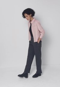Woman with curly hair wearing a pink shirt, black top, gray pants, and black shoes standing with hands in pockets, looking down.