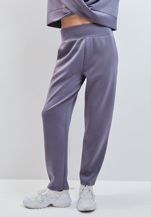 Personne portant un pantalon de jogging lavande et des baskets blanches épaisses, debout devant un fond clair uni.