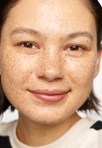 Gros plan sur un visage avec une peau brun clair, de nombreuses taches de rousseur et des cheveux noirs et raides. Les lèvres sont d'un rose doux ; les yeux sont marron. Maquillage minimal visible.