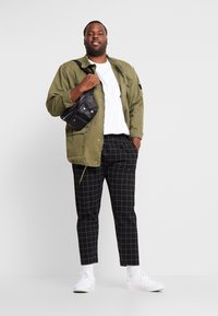 Olijfgroene jas, witte t-shirt, zwart-wit geblokte broek en witte sneakers. Houdt een zwarte crossbodytas vast met meerdere vakken.