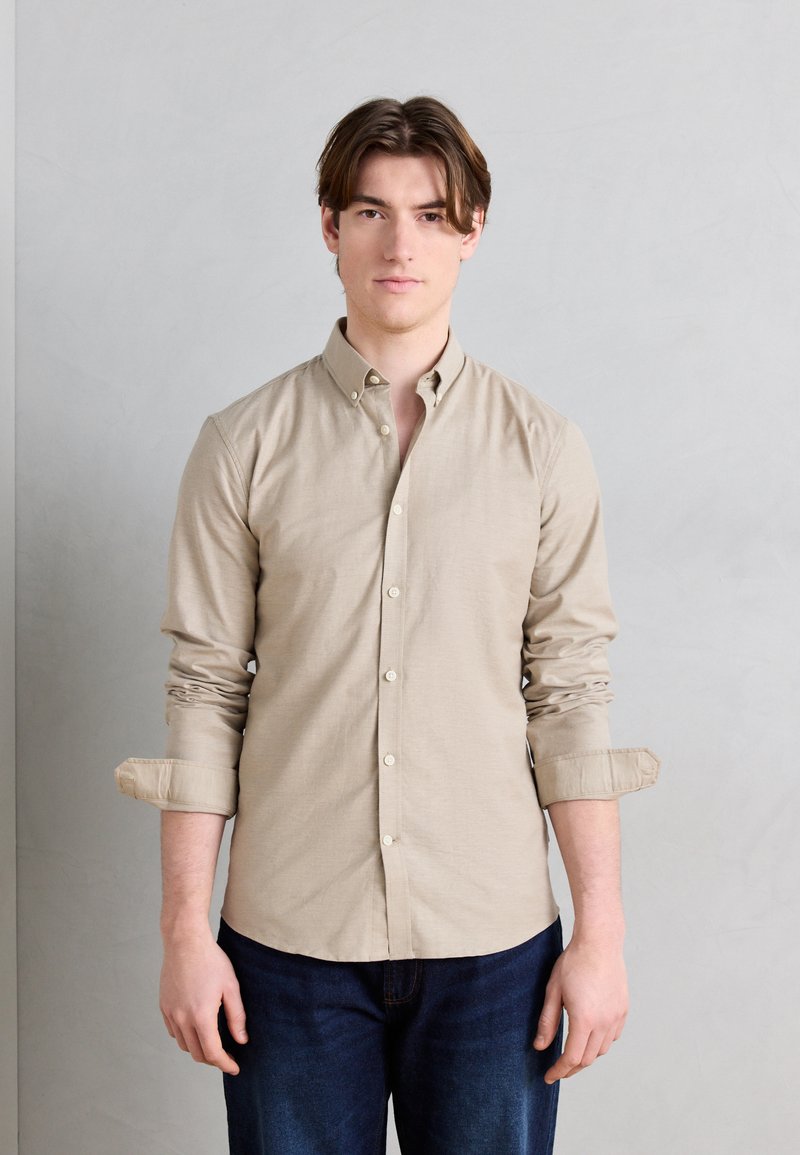 Νέος άνδρας με καστανά μαλλιά, φορώντας μπεζ πουκάμισο με κουμπιά και γυρισμένα μανίκια, καθώς και σκούρο μπλε τζιν, που στέκεται μπροστά σε απλό ανοιχτογκρι φόντο.