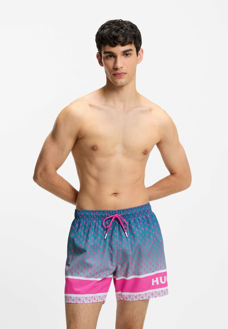 Jeune homme debout torse nu portant un short de bain bleu et rose à motifs avec un cordon rose sur fond blanc.