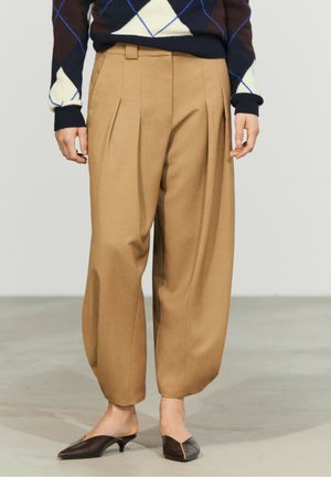 Personne portant un pantalon ample beige, des escarpins pointus foncés et un pull à losanges foncé avec des motifs blancs et bleus.