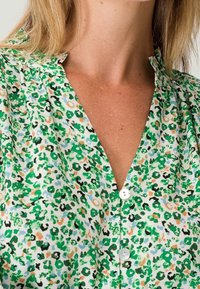 Blusa verde y blanca con un diseño de patrones, con escote en V, que presenta una combinación de flores y estampado de leopardo, confeccionada en tela ligera y con cierre de botones en la parte delantera.